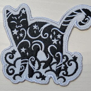 May include: Black and white embroidered iron-on patch of a cat with a swirling design and stars. The cat is outlined in white and the background is white.