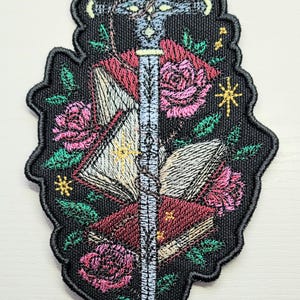 May include: An embroidered patch with a sword piercing through a stack of books and roses. The background is black with gold stars and green leaves.