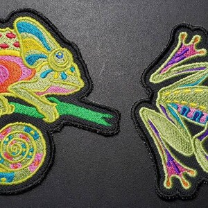May include: Embroidered patches featuring a chameleon and a frog. The chameleon patch is multi-colored, with pink, orange, yellow, and blue details. The frog patch is green with red eyes and purple accents. Both patches have a black border.