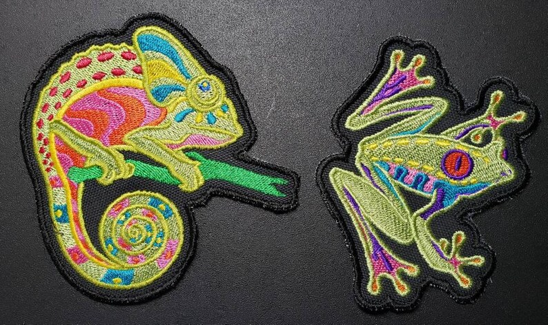 May include: Embroidered patches featuring a chameleon and a frog. The chameleon patch is multi-colored, with pink, orange, yellow, and blue details. The frog patch is green with red eyes and purple accents. Both patches have a black border.