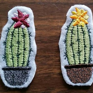 May include: Two embroidered cactus patches with different colored flowers. The cactus on the left has a pink and red flower, while the cactus on the right has a yellow flower. Both cacti are green and in brown pots.
