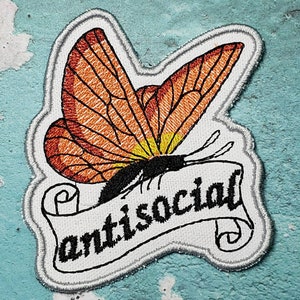 May include: An embroidered iron-on patch featuring an orange butterfly with black wings and a yellow body. The butterfly is perched on a white scroll with the text "antisocial" in black.