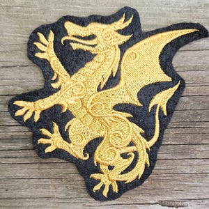 May include: An embroidered patch featuring a gold dragon silhouette on a black background. The dragon is depicted in a side profile with intricate stitched details, including a curved tail, outstretched wings, and clawed feet, all rendered in metallic gold thread.