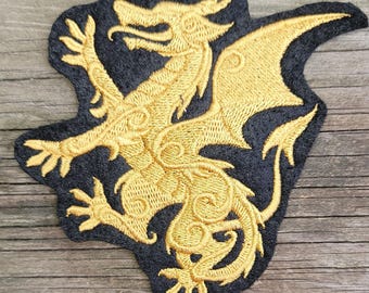 May include: An embroidered patch featuring a gold dragon silhouette on a black background. The dragon is depicted in a side profile with intricate stitched details, including a curved tail, outstretched wings, and clawed feet, all rendered in metallic gold thread.