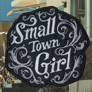 May include: A black embroidered patch with white stitching. The patch features a decorative design with the text "Small Town Girl".