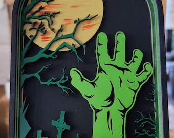 May include: A layered wooden Halloween decoration showing a green zombie hand emerging from a grave. The background features a full moon, trees, and tombstones, set against a black backdrop. The artwork is framed in a dark arch.