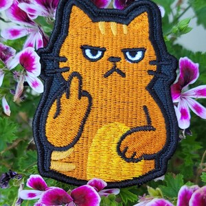 May include: Embroidered patch featuring an orange cat with a black outline, giving the middle finger. The cat has blue eyes and a grumpy expression. The patch is surrounded by pink and purple flowers.