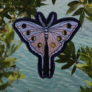 Op de afbeelding: Geborduurde vlinderpatch met een gedetailleerd ontwerp. De patch heeft een lichtblauwe en roze vlinder met zwarte en gouden accenten. De patch is omlijnd in donkerblauw en zwart.