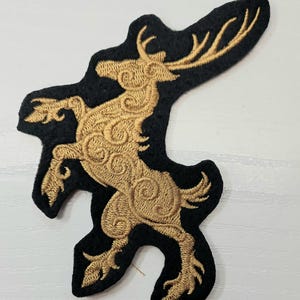 May include: A black fabric patch featuring an embroidered gold thread design of a rearing stag. The deer is depicted in a stylized, swirling pattern with prominent antlers, captured in a dynamic pose on a dark, felt-like material background.
