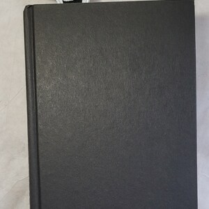 May include: A black hardcover book with a red, black, and white embroidered bookmark. The bookmark features a stylized design. The book is closed and has a textured cover.