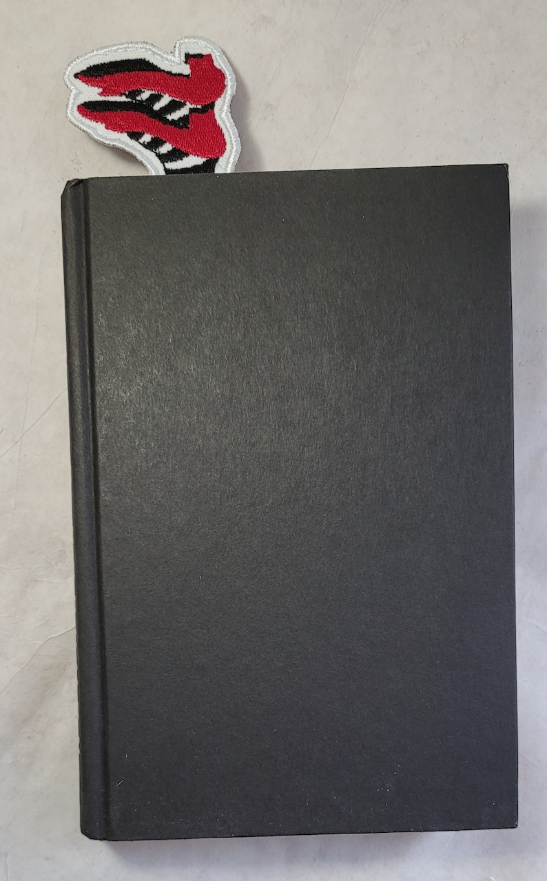 May include: A black hardcover book with a red, black, and white embroidered bookmark. The bookmark features a stylized design. The book is closed and has a textured cover.