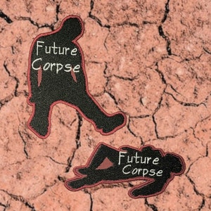 May include: Two black embroidered patches with red stitching. The patches are shaped like silhouettes of people. One patch shows a person standing with the text "Future Corpse" in white. The other patch shows a person lying down with the same text.