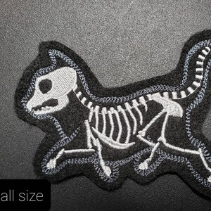 K&ouml;nnte beinhalten: Schwarzer Filz-Patch mit einem wei&szlig;en gestickten Skelett einer Katze. Die Katze rennt und hat einen langen Schwanz.