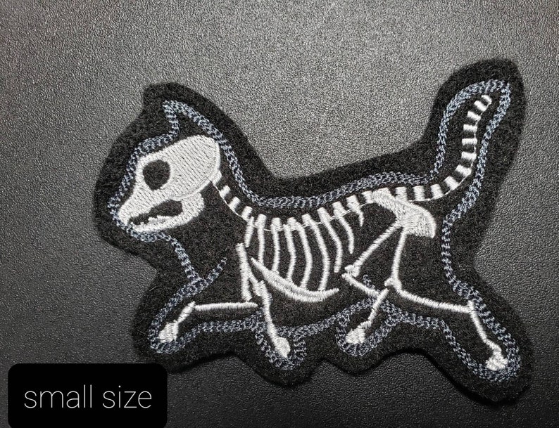 K&ouml;nnte beinhalten: Schwarzer Filz-Patch mit einem wei&szlig;en gestickten Skelett einer Katze. Die Katze rennt und hat einen langen Schwanz.