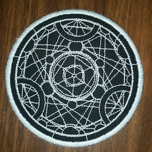 May include: A black and white embroidered patch with a circular design. The design features a complex pattern of overlapping circles and lines.