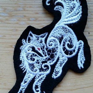 May include: White embroidered wolf design on a black felt patch. The wolf is depicted in a stylized, intricate design with swirling lines.