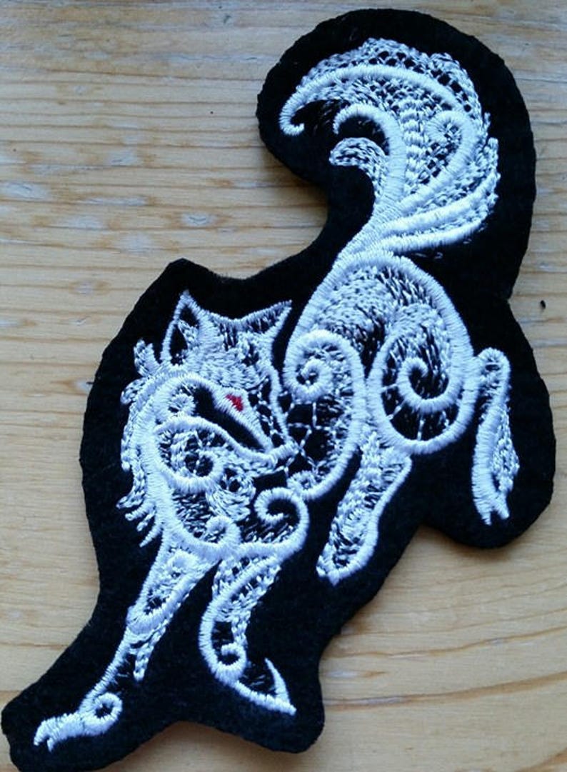 May include: White embroidered wolf design on a black felt patch. The wolf is depicted in a stylized, intricate design with swirling lines.