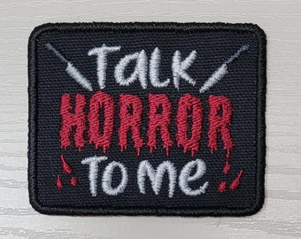 Könnte beinhalten: Schwarzer gestickter Aufnäher mit weißen und roten Nähten. Der Aufnäher sagt "Talk Horror To Me" mit zwei stilisierten Messern auf jeder Seite des Textes.