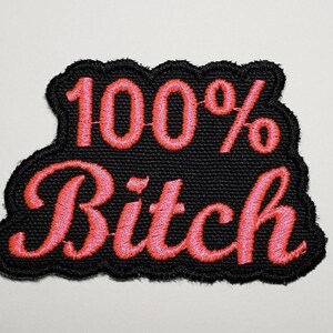 May include: Embroidered patch with the text "100% Bitch" in pink script. The text is stitched onto a black fabric with a scalloped edge. The patch is designed to be ironed or sewn onto clothing or accessories.