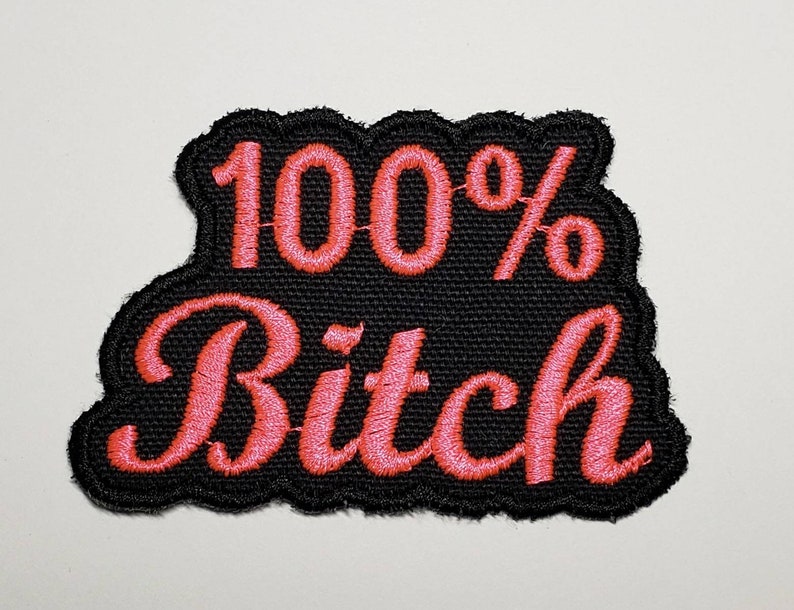 May include: Embroidered patch with the text "100% Bitch" in pink script. The text is stitched onto a black fabric with a scalloped edge. The patch is designed to be ironed or sewn onto clothing or accessories.