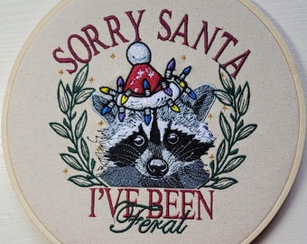 Könnte beinhalten: Gesticktes Stickrahmen-Kunstwerk mit einem Waschbären, der einen Weihnachtsmannhut und eine Lichterkette trägt. Der Text lautet "SORRY SANTA I'VE BEEN FERAL" in bordeauxroter und grüner Stickerei. Der Waschbär ist von einem grünen Blätterkranz umgeben.