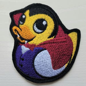 May include: Embroidered patch featuring a yellow duck with a vampire theme. The duck has a black cape, purple vest, and a small fang. The patch has a black border.