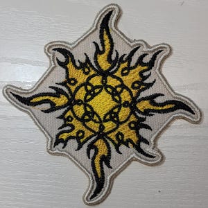May include: A white embroidered patch with a yellow and black sun design. The sun has a circular center with a black and yellow design and is surrounded by yellow flames.