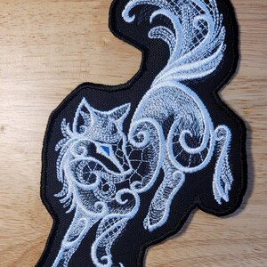 May include: White embroidered wolf design on a black fabric patch. The wolf is depicted in a stylized, intricate pattern with a swirling, lace-like design.