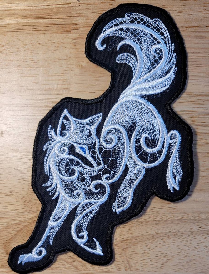 May include: White embroidered wolf design on a black fabric patch. The wolf is depicted in a stylized, intricate pattern with a swirling, lace-like design.