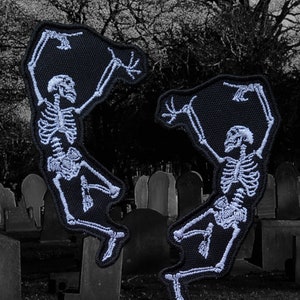 May include: Two white embroidered skeleton patches on a black fabric background. The skeletons are in a dancing pose with their arms raised above their heads. The patches are set against a blurry background of a graveyard.