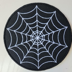 May include: Round black patch featuring a white spider web design. The web is intricately detailed, radiating from a central point. The patch has a black border and is likely made of fabric.