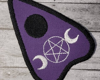 May include: Purple fabric patch with a black circle, a white pentagram, and three white crescent moons. The patch is shaped like a teardrop and has a black border.