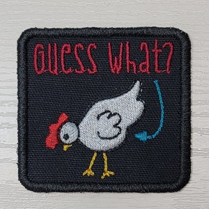 May include: Embroidered patch with a black background and a white chicken illustration. The text "GUESS WHAT?" is embroidered in red above the chicken. A blue arrow points towards the chicken's rear.