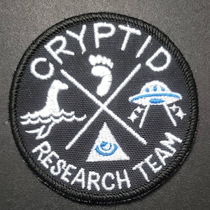 May include: A black embroidered patch with white stitching. The patch features the words "Cryptid Research Team" in a circle around a white triangle with a blue eye inside. The triangle is crossed by two white sticks, one with a footprint, one with a sea monster, and one with a UFO.