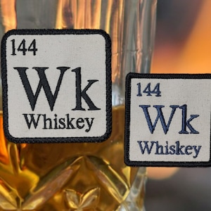 May include: Two beige fabric patches with black borders. Each patch has a white background with black text. The text on the patches reads "144 Wk Whiskey". The patch on the left has black lettering, and the patch on the right has blue lettering.