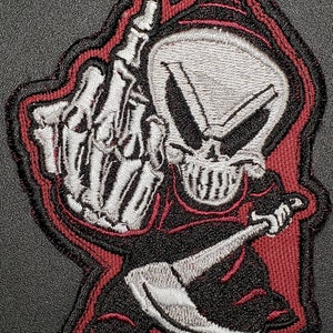 May include: Embroidered patch featuring a grim reaper figure giving the middle finger. The design includes a white skeleton hand, a skull, and a black robe with red accents. The patch is outlined in black and red.
