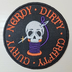 May include: Round black patch with orange text that reads "Nerdy Dirty Curvy Crafty." A white skull with a needle and thread is in the center, above a purple and orange spool of thread. The patch is a fun, quirky item.
