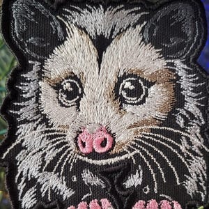 May include: Embroidered patch featuring a detailed opossum face. The patch has a black border and background, with the opossum's face in shades of gray, brown, and pink. The opossum's eyes are large and round, and its nose is pink.