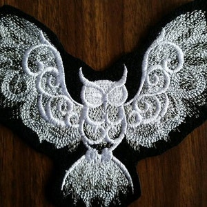 May include: White embroidered owl patch with intricate wing designs on a black fabric background.