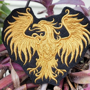 May include: A black fabric patch featuring an intricate gold embroidered phoenix with wings spread wide. The design showcases detailed stitching of the bird's feathers, head, and talons, creating a bold contrast against the dark background fabric.