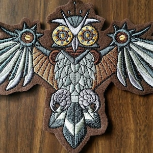May include: A brown felt patch with an embroidered owl with large, round, gold eyes. The owl has brown and gray wings with a silver and white design. The owl is holding a silver and white feather in its talons.