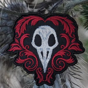 May include: Embroidered patch featuring a white skull with a beak, surrounded by red floral designs on a black background.