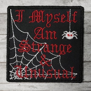 May include: Black embroidered patch with a spider web design and the text "I Myself Am Strange & Unusual" in red lettering. A red spider with white eyes hangs from the web.
