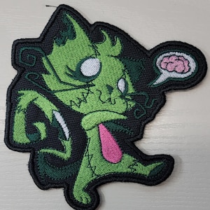 May include: An embroidered patch featuring a stylized green cat with stitched details, large white eyes, and a pink chest patch. The cat is depicted with a thought bubble containing a pink brain, all outlined in black thread on a black background.