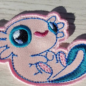May include: Embroidered patch featuring a cartoon axolotl in shades of pink, teal, and blue. The axolotl has large, expressive eyes and a smiling expression. The patch is outlined in pink and blue stitching.