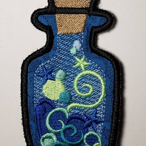 May include: Embroidered patch in the shape of a bottle, featuring a blue denim background. Inside, there are embroidered elements of sea life, including a green swirl, bubbles, and starfish. The bottle has a cork top and a black border.