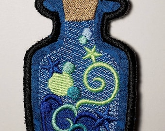 May include: Embroidered patch in the shape of a bottle, featuring a blue denim background. Inside, there are embroidered elements of sea life, including a green swirl, bubbles, and starfish. The bottle has a cork top and a black border.