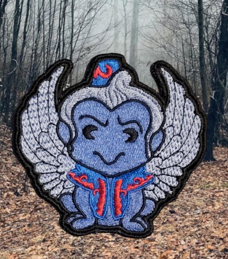 Puede incluir: Parche bordado de un mono azul alado con un turbante rojo y azul. El mono tiene una expresi&oacute;n traviesa y lleva una t&uacute;nica azul y roja.