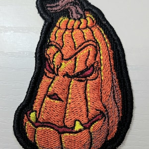 K&ouml;nnte beinhalten: Ein gestickter Aufn&auml;her in Form eines l&auml;nglichen, orangefarbenen K&uuml;rbisses mit einem grimmigen, geschnitzten Gesicht. Der K&uuml;rbis hat tiefe vertikale Rillen, einen dunklen Stiel und einen dicken schwarzen Rand. Das Gesicht zeigt schmale Augen.