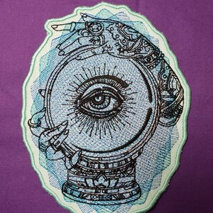 May include: An embroidered patch featuring a hand holding a crystal ball with an eye inside. The hand is adorned with intricate details and the crystal ball is surrounded by a blue and green watercolor-like design.